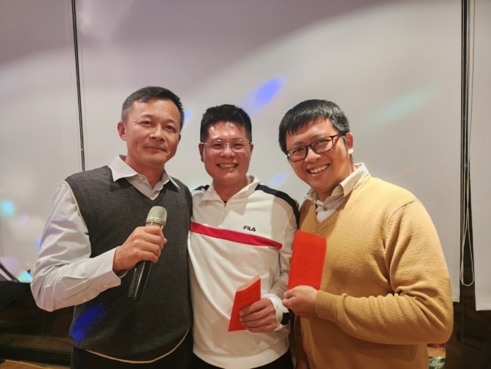 A group of three men standing together and smiling. One is holding a microphone and two are holding red envelopes.