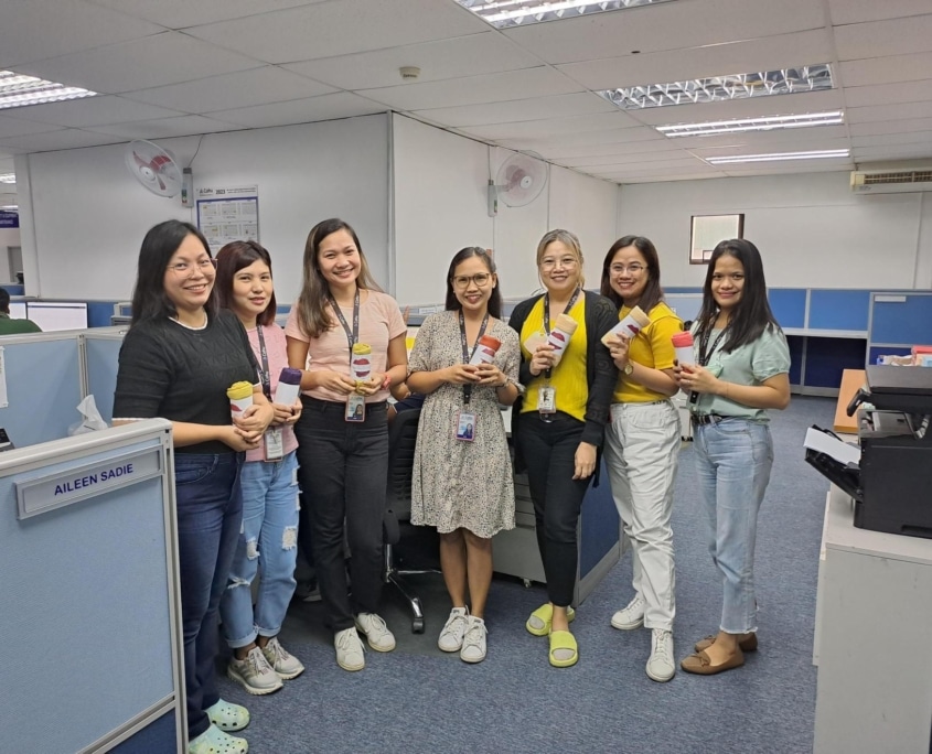 A group of seven women smiling and each holding a rolled up shirt. They are in an office and standing outside of a cubicle.