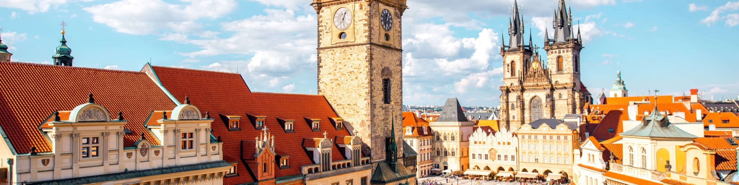 A landscape showing a European city with orange rooftops, a brick clocktower and a gothic style church in the background. A small town square can be seen at the bottom.