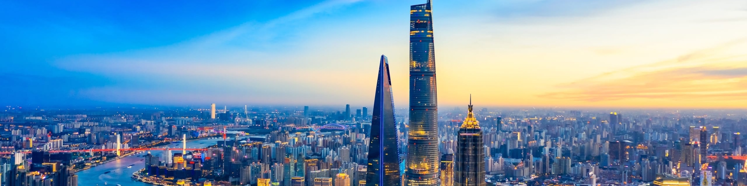 A modern cityscape with hundreds of skyscrapers as the sun is just barley starting to set. There are three steel pillar shaped buildings in the foreground that are very sleek and modern looking.