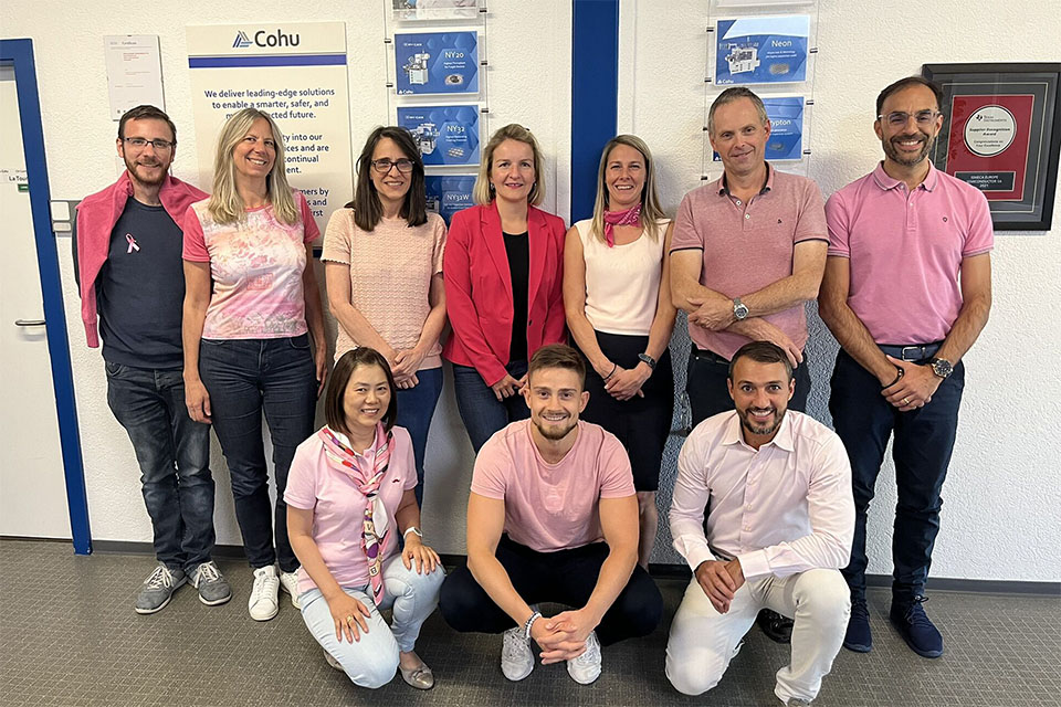 A group of 10 people together, all wearing various shades of pink shirts. They are in front of a white wall adorned with pictures of Cohu products.