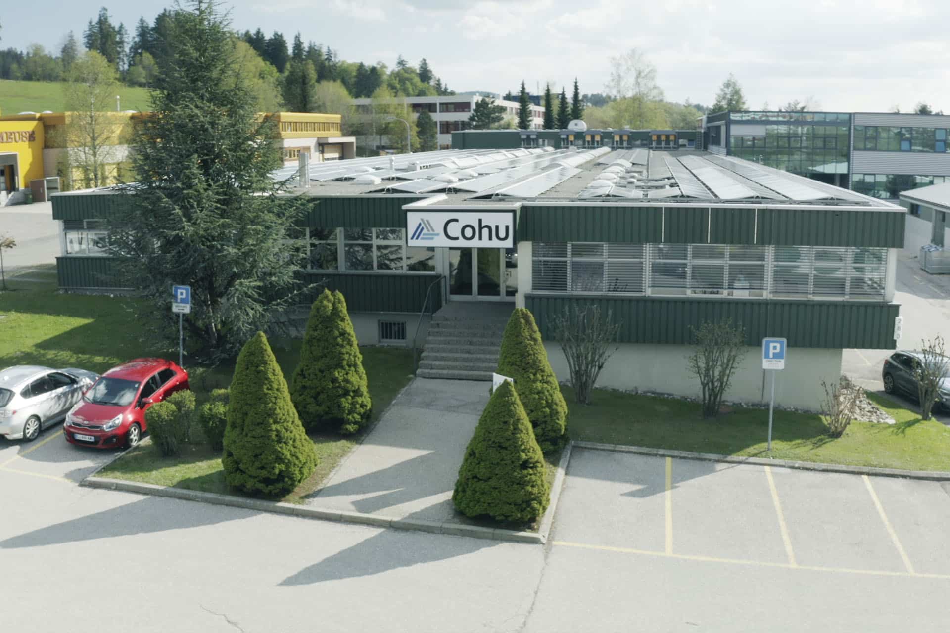 An aerial image of a one story building, lined with windows and with a sign that reads Cohu. There is a defined, paved path to enter lined with shrubs and cars in the foreground of the parking lot.