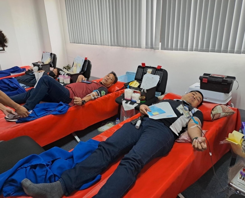 Three men laying down on red colored beds. They have IVs in their arms and are giving blood next to boxes of medical supplies.