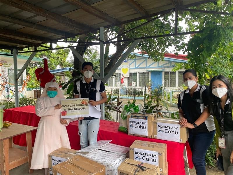 A group of 4 people standing under a covered roof looking at the camera. They are all wearing masks and standing next to 9 boxes that read Donated by Cohu.