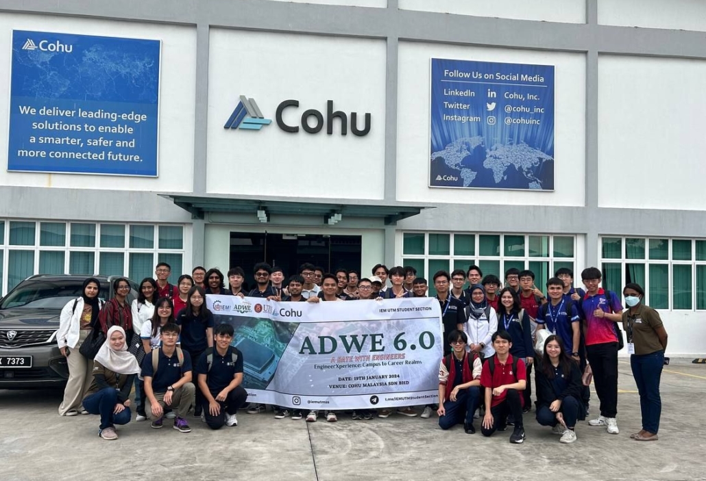 A group of 45 people standing in front of an entrance to a building with a sign that reads Cohu. They are holding a large banner that reads ADWE 6.0 A Date with Engineers.