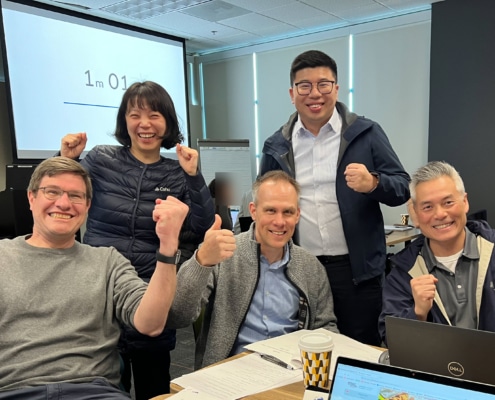 A group of five people smiling and holding their fists up in excitement. Three are sitting at a table and two are standing and there is a projector screen behind them that reads 1m 01.