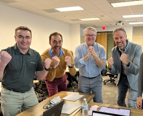 A group of four men smiling and excited, holding their fists up to their chest. They are standing in front of an office table in an office environment.