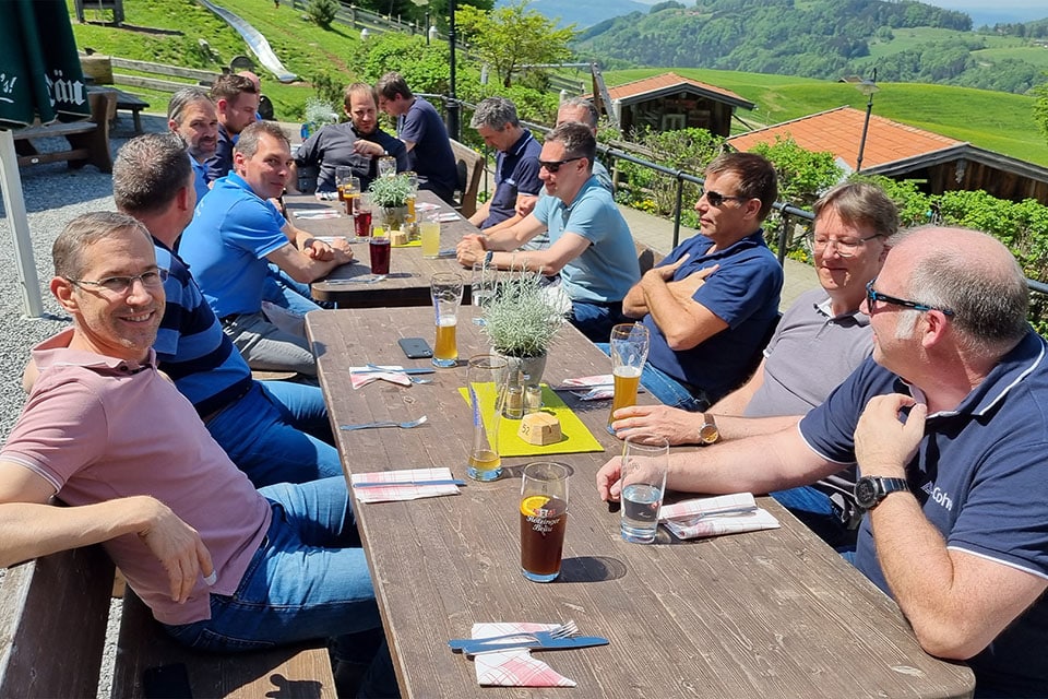 A group of 14 men sitting together outsde at a picnic table with beer and silverware on the table. Only one man is smiling and looking at the camera while the rest are in conversation with one another.