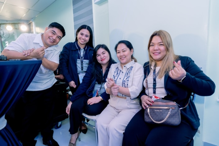 A group of 5 people sitting together and smiling. Three are holding their fingers up to make a heart sign.