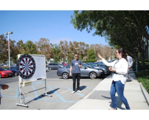 Three people standing in a parking lot throwing darts at a dartboard that is attached to a rolling whiteboard. There are rows of cars in the background.