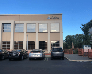 A two story tan building with windows and a sign that reads Cohu. There are four cars parked in front of the building.