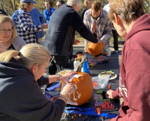 Approximately 11 people outside watching two groups of people carve pumpkins. One group is using a stencil and a carving kit.