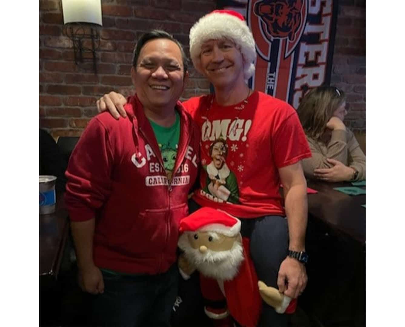 Two men standing together smiling wearing. One man is wearing red and green and the other is wearing a Santa hat and a Christmas sweater.