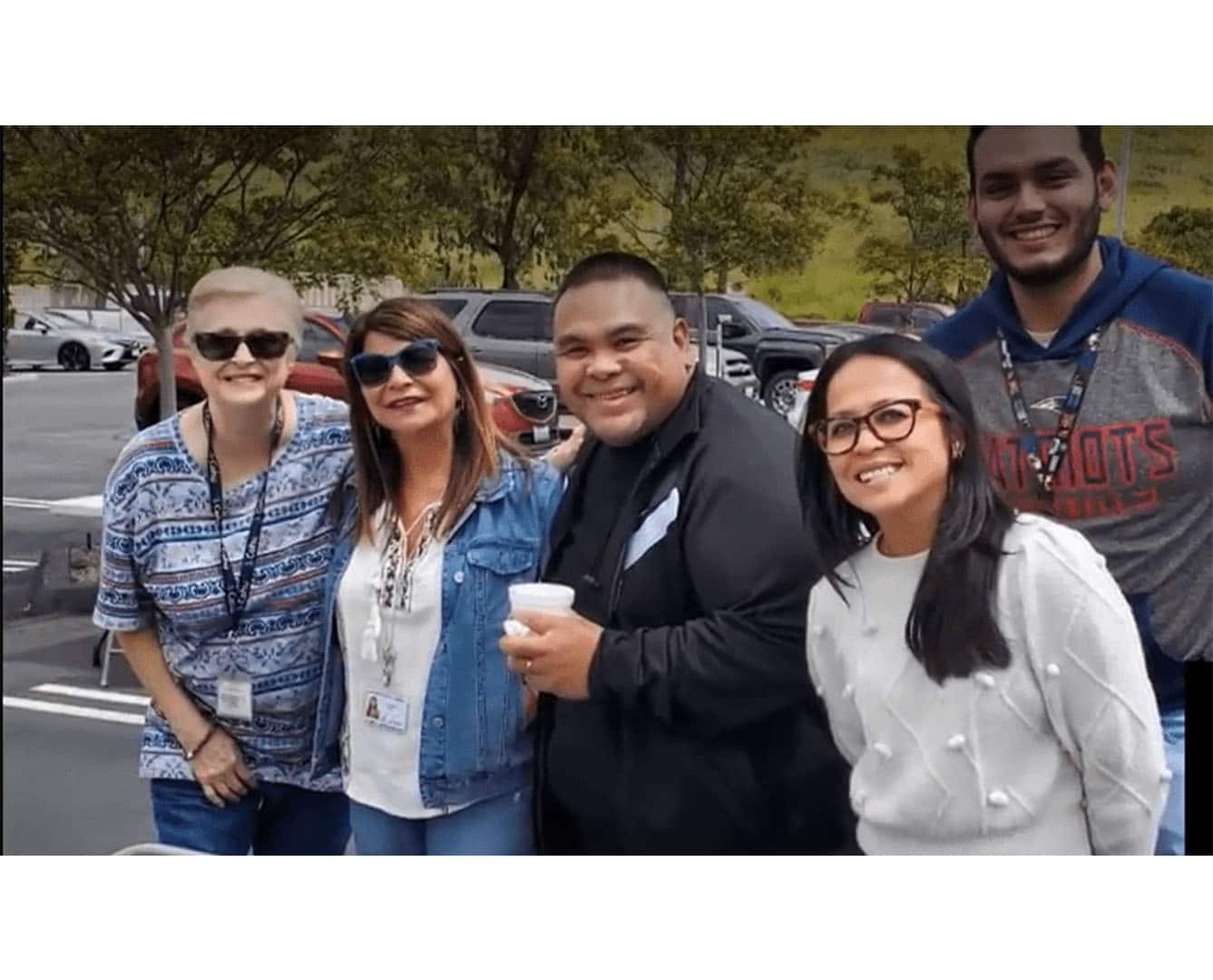 Three women and two men standing together smiling in a parking lot. Two of the women are wearing sunglasses.