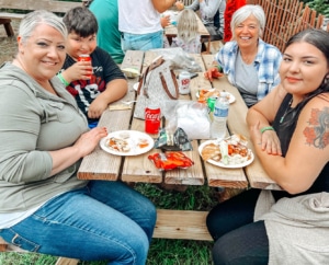 A group of 4 people sitting together at a picnic table smiling with food and drinks on the table in front of them.