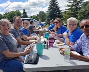 A group of 6 people sitting at a picnic table smiling with food and drinks on the table. There is a group of three at another table in the background.