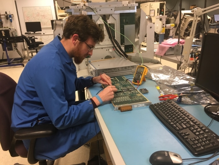 A man working on an electrical circuit board at a desk indoors. He is wearing a blue coat and is surrounded by other machinery.