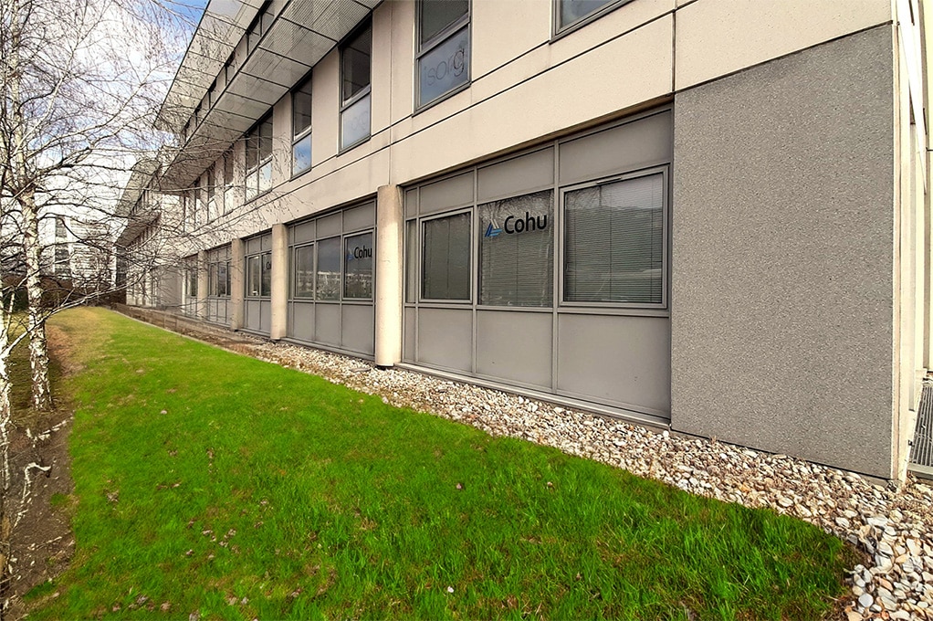 The side of a three story building, lined with pebbles and green grass. The building has many windows and Cohu visible on four of the windows.