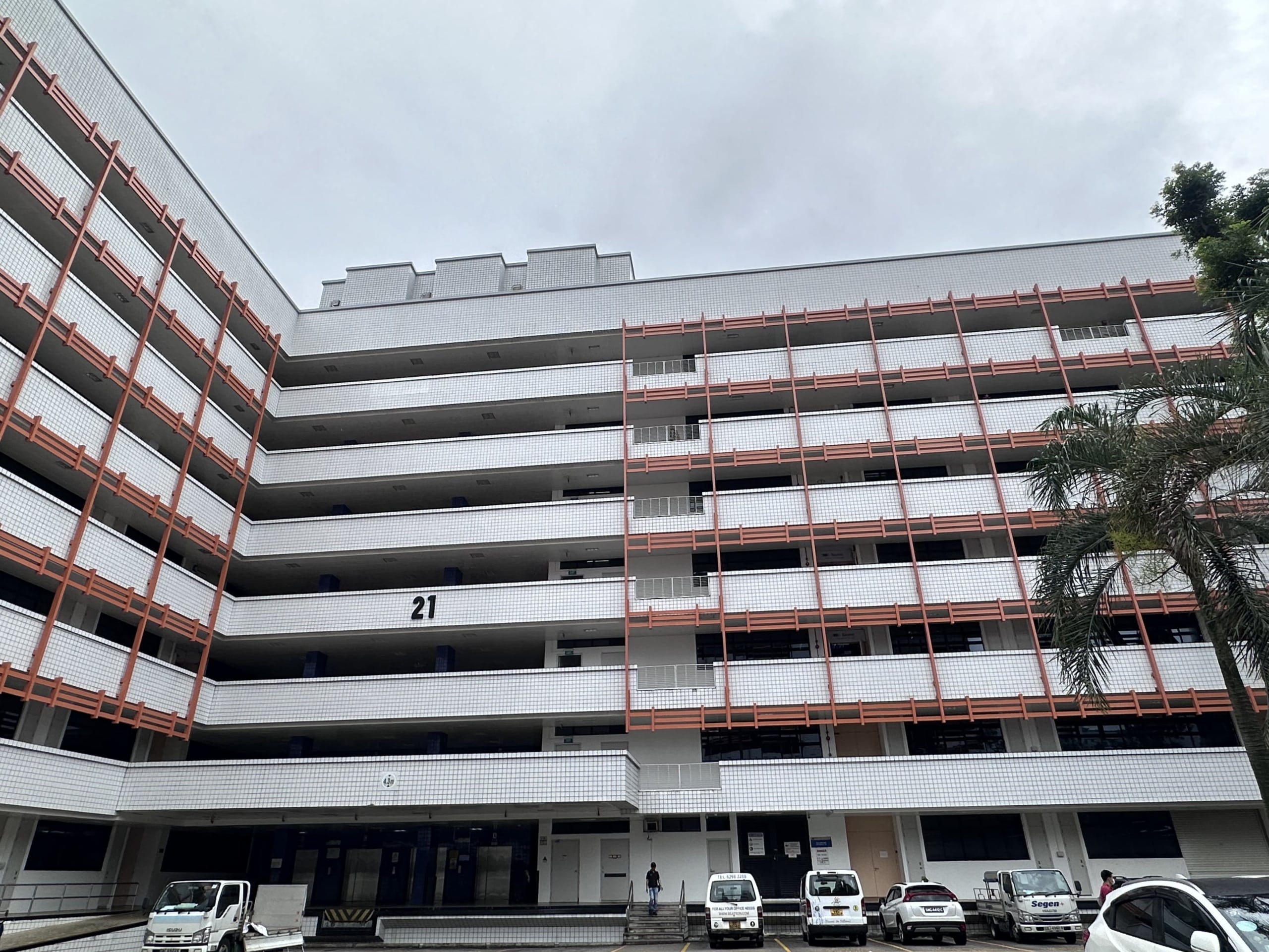 A long seven story building with red and white accents on a cloudy day. There are at least six cars in the parking lot in front of the building.