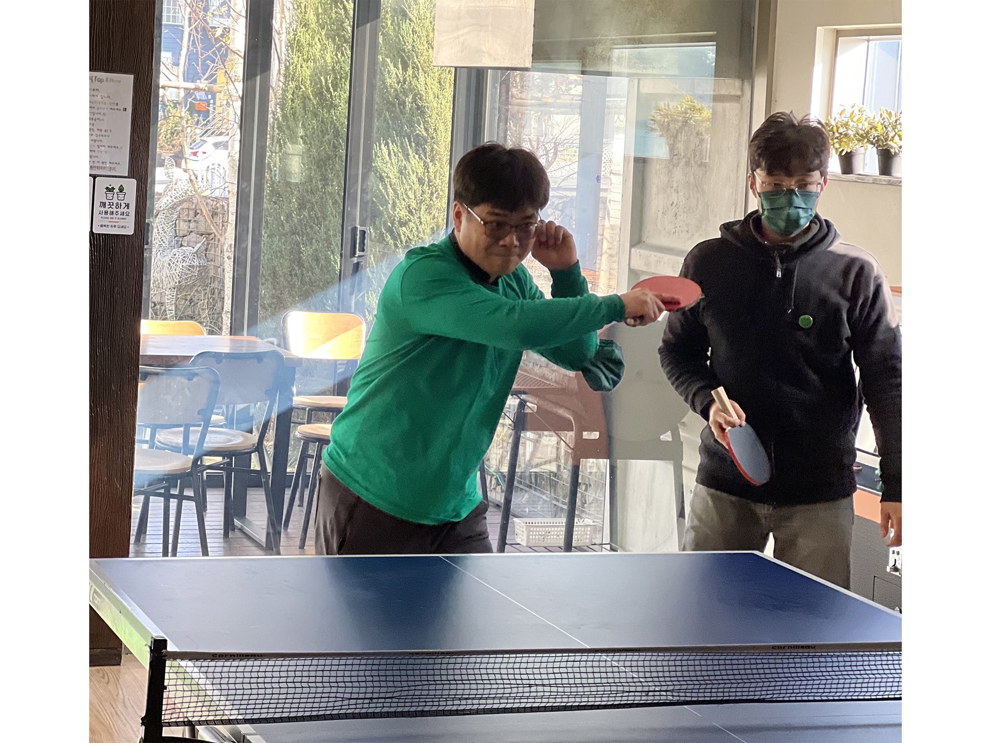 Two men play ping pong indoors at a table. One man is wearing a mask and there are windows in the background.