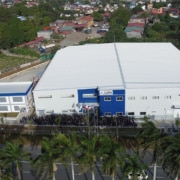 An aerial image of a large two story building that is white and blue in color with a smaller, square two story building to the left. There are smaller, residential houses in the background and a group of people and palm trees in the foreground.