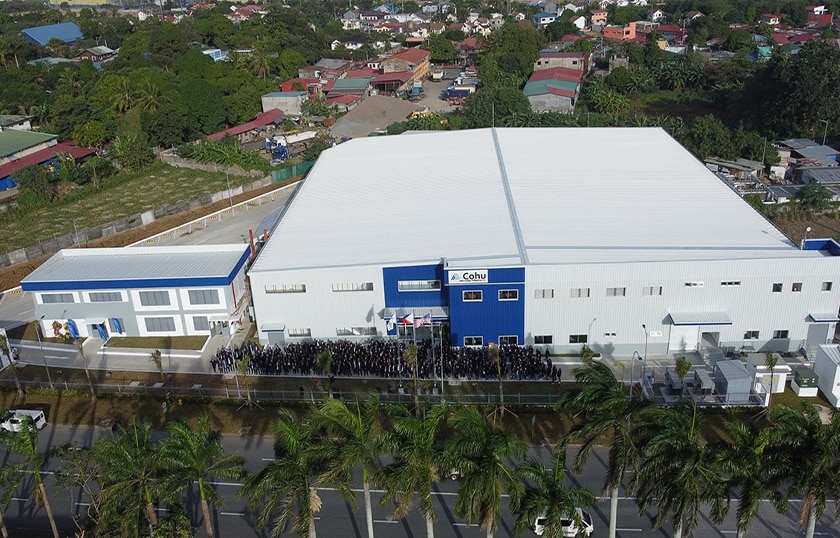 An aerial image of a large two story building that is white and blue in color with a smaller, square two story building to the left. There are smaller, residential houses in the background and a group of people and palm trees in the foreground.