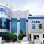 A building comprised of three distinct sections - one cylindrical, one a taller rectangle, and a smaller square one with Cohu on the front. The buildings are all shades of grey and blue and reflective with three cars parked in front of the buildings.