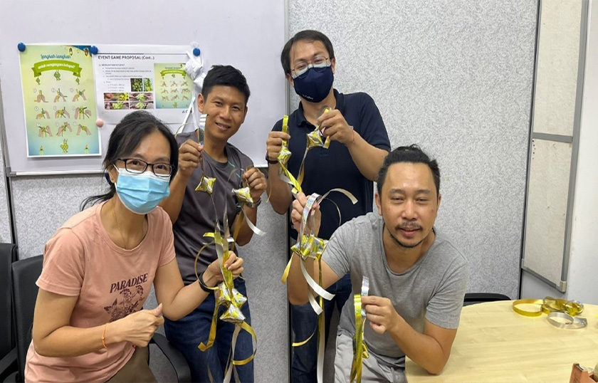 Three men and one woman in an office space holding up golden ribbon and smiling. One man and one woman are wearing masks.