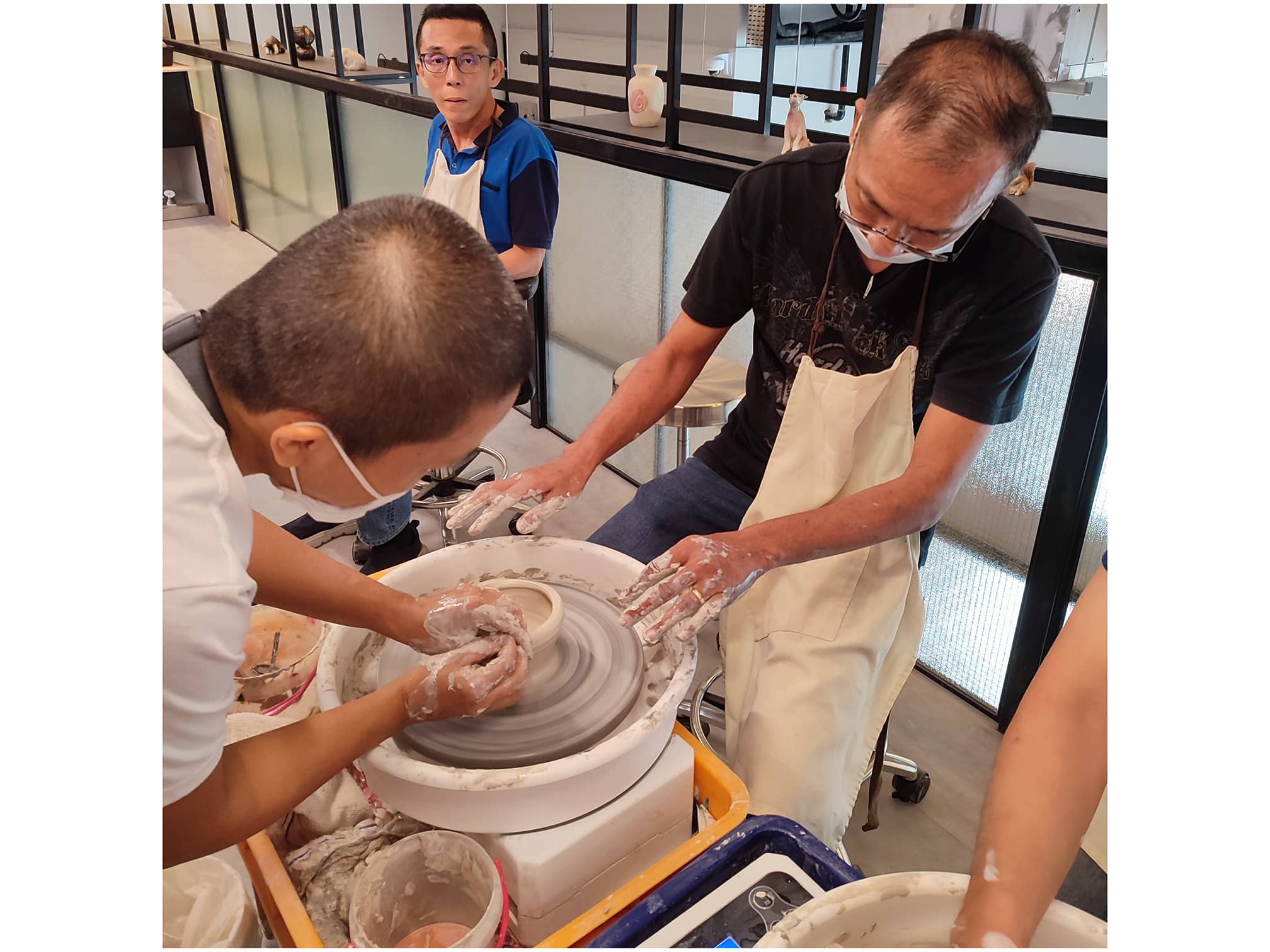 Two men sitting at a pottery wheel working on a piece of clay while a man looks on in the background.