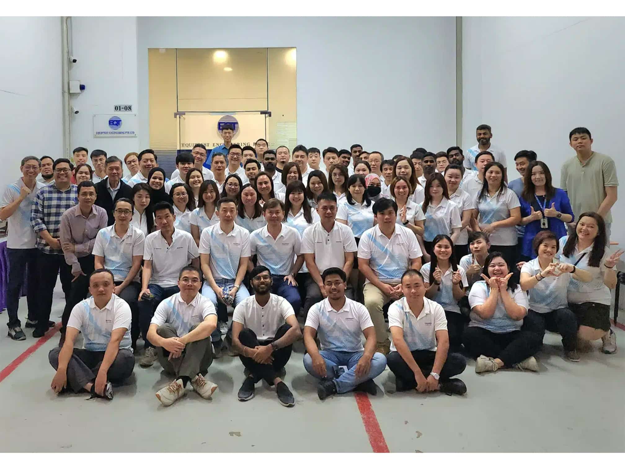 A group of about 80 men and women smiling for a picture in a tall room with white walls and EQT written on the back wall.