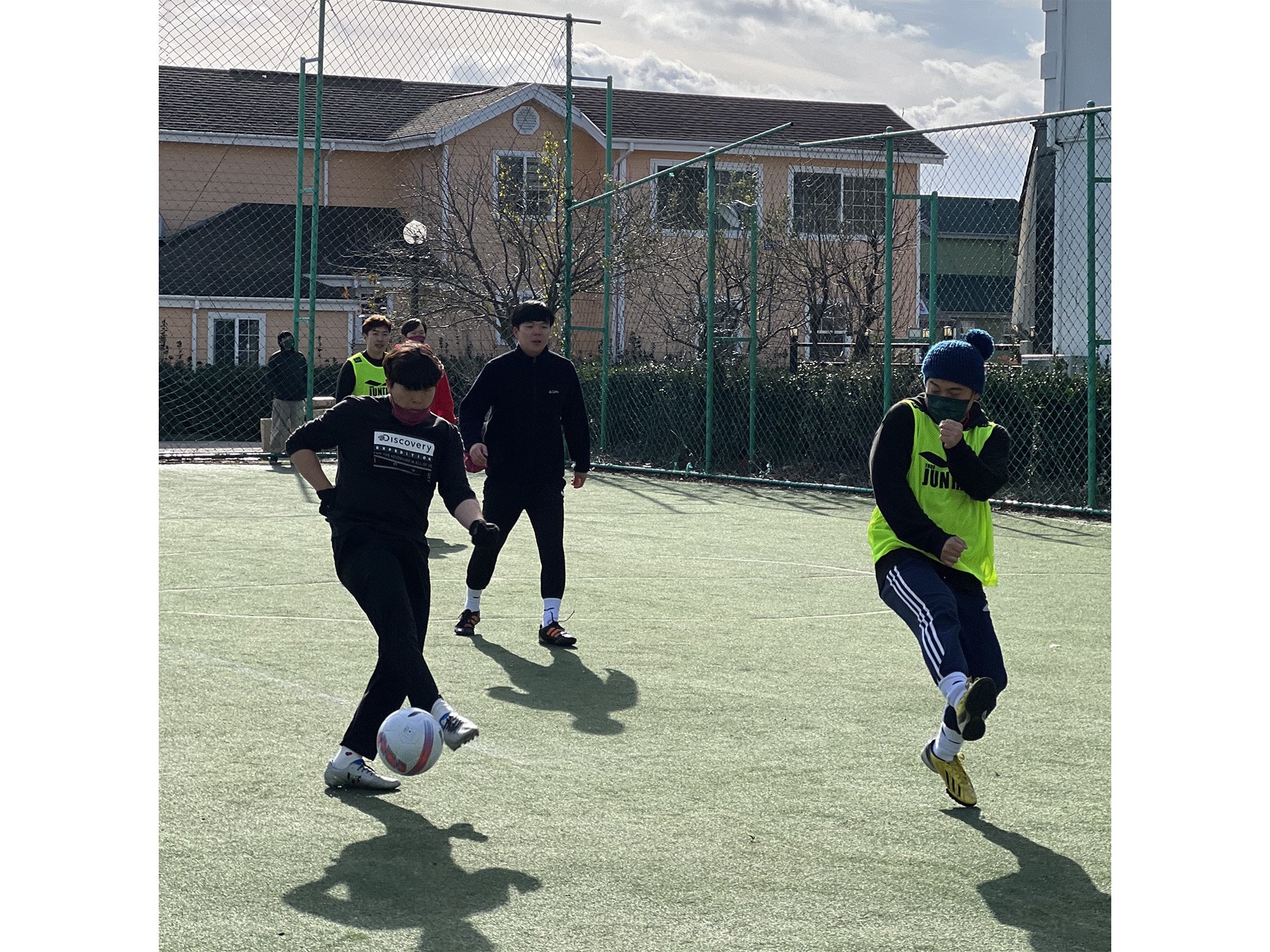 Five men on a turf field while one man has the soccer ball at his feet. The turf is enclosed by a fence and an orange building is in the background.