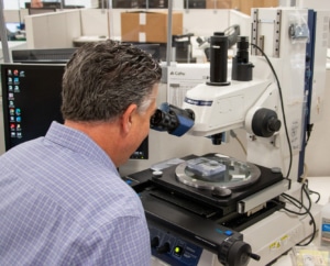 A man looks through a microscope with his back to the camera. There is a blank computer screen in the background.