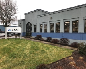 The outside of a one story building with a glass entryway, nine windows, and a sign that reads Cohu 6 Court Drive. There is a short lawn, sidewalk, and shrubs bordering the building.