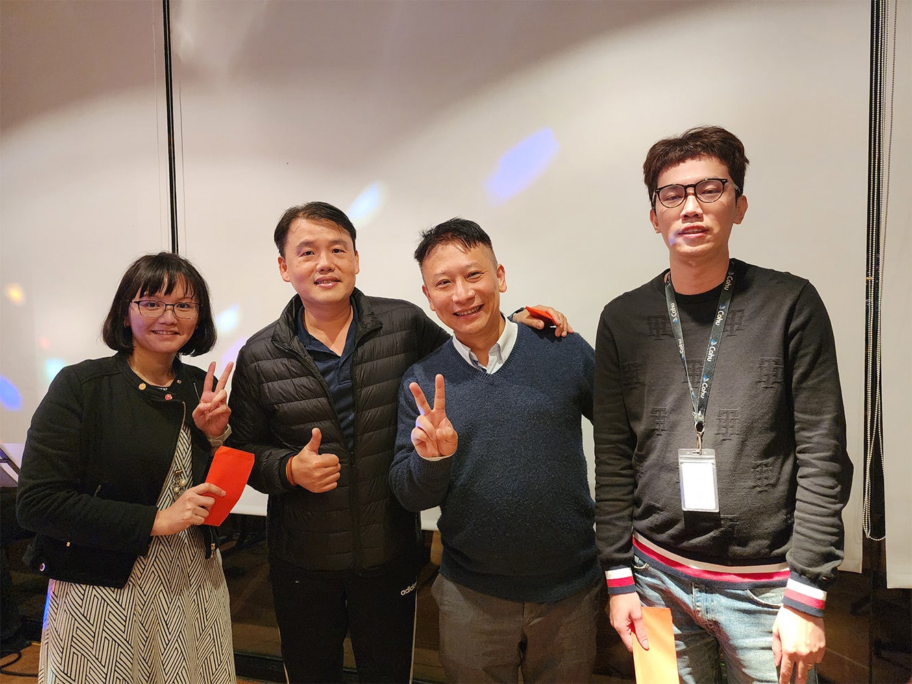 Three men and one woman standing together smiling with two people making the peace sign with their hands.